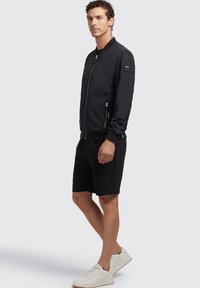 Jachetă bomber neagră cu închidere cu fermoar, buzunare laterale și mansete ribbed. Asortată cu pantaloni scurți negri și adidași albi. Textură fină.