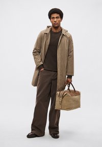 Bézs esőkabát, barna kötött pulóver és barna bő szárú nadrág. Szürke táska barna díszítéssel. A modell rövid, göndör hajú, karjai lazán állnak.