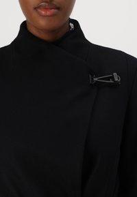 Cappotto di lana nero con colletto alto, dotato di un grande gancio nero sulla spalla. Texture liscia con linee pulite e senza modelli visibili.
