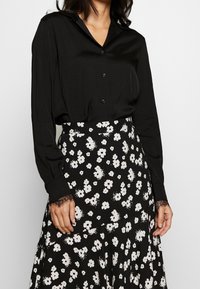 Mujer vistiendo una blusa negra abotonada con puños de encaje, metida en una falda negra con estampado floral blanco.