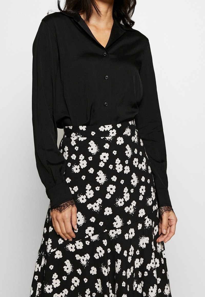 Mujer vistiendo una blusa negra abotonada con puños de encaje, metida en una falda negra con estampado floral blanco.