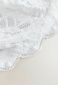 Fehér csipke anyag virág- és geometriai mintákkal, hullámos szegélyekkel és finom hálós textúrával világos háttéren.