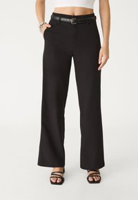Schwarze weite Hose mit Gürtel, die eine glatte Textur, flachen Front und schräge Seitentaschen aufweist. Kombiniert mit schwarzen Sandalen mit Riemen.