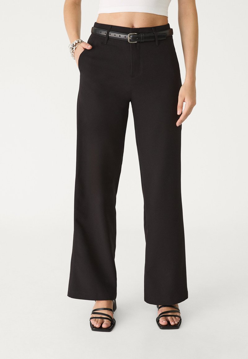 Schwarze weite Hose mit Gürtel, die eine glatte Textur, flachen Front und schräge Seitentaschen aufweist. Kombiniert mit schwarzen Sandalen mit Riemen.