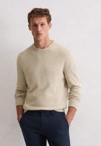 Maglione beige a maglia con superficie texture, scollo rotondo e polsini a costine, abbinato a pantaloni blu scuro.