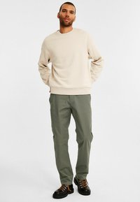 Beiger Sweatshirt mit Rundhalsausschnitt, kombiniert mit olivgrünen Chinos und schwarzen strukturierten Stiefeln mit orangefarbenen Schnürsenkeln. Einfaches Design, lässige Passform.