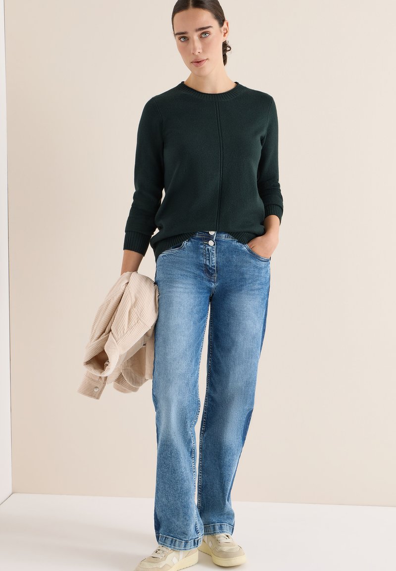 Maglione lavorato a maglia verde scuro con scollatura rotonda, abbinato a jeans larghi blu. Giacca in corduroy beige appoggiata su un braccio.
