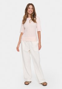 Šviesiai rožinė mezgimo palaidinė su trumpomis, raukšlėtomis rankovėmis ir tekstūruotu raštu, derinama su laisvai krentančiomis baltomis kelnių ir kasdieniais šlepetėmis.