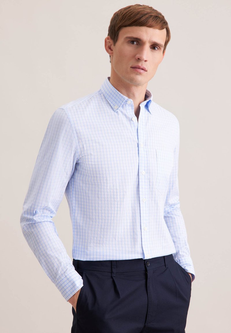 Junger Mann mit kurzem braunem Haar, der ein hellblau kariertes Hemd mit Knöpfen und dunkle Hose trägt, steht mit den Händen in den Taschen.