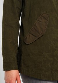 Giacca verde scuro con un tessuto testurizzato e una tasca imbottita angolata. La manica ha un polsino cucito, che aggiunge particolare al design.