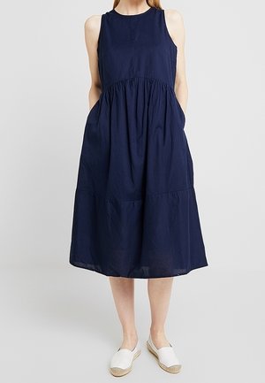 Robe midi bleu marine sans manches avec taille froncée et poches, portée avec des espadrilles blanches slip-on sur un fond gris clair.