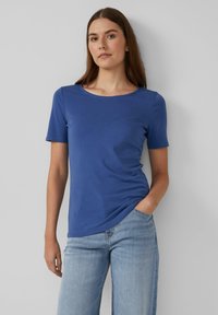 Młoda kobieta z długimi brązowymi włosami, ubrana w zwykłą niebieską koszulę z krótkim rękawem i jasnoniebieskie dżinsy, stojąca na białym tle.