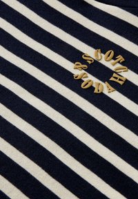 T-paita, jossa on vaaleansinisiä ja kerma-valkoisia vaakasuoria raitoja, esittelee pyöreän beige brodeeratun logon, jossa lukee "Scotch & Soda." Pehmeä rakenne.