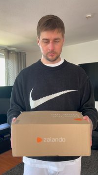 Pruun pappkast, millel on oranž "zalando" logo, mida hoiab inimene mustas svetrises, millel on valge Nike logo, seistes siseruumides.