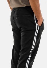 Zwarte trainingsbroek met twee witte verticale strepen aan de zijkanten, een achterzak met ritssluiting en een logo-opdruk op het linkerbeen.