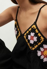 Robe noire avec un décolleté en V ornée de motifs floraux brodés colorés en rose, orange et jaune sur un fond texturé. Fines bretelles.