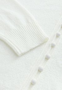 Cardigan blanc en tricot avec une finition texturée, doté d'une manche côtelée et d'un tissu doux, agrémenté de boutons ronds blancs sur le devant.