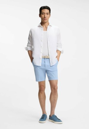 Hombre de pie con las manos en los bolsillos, vistiendo una camisa blanca abotonada, camiseta interior blanca, pantalones cortos azul claro, cinturón beige y zapatillas azules.