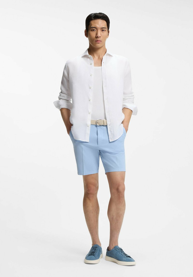 Uomo in piedi con le mani nelle tasche che indossa una camicia bianca con bottoni, una maglietta bianca sotto, pantaloncini azzurri, cintura beige e scarpe da ginnastica blu.