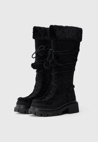 Cizme negre din piele întoarsă cu înveliș moale, design cu șireturi, pompoane decorative, talpă groasă de cauciuc și accente de blană în jurul părții superioare.