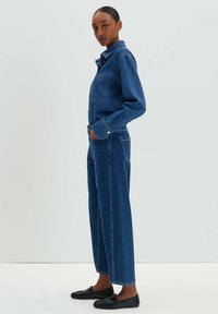 Denim-Overall mit Knopfverschluss vorne, langen Ärmeln und weitem Beinschnitt. Dunkelblaue Farbe mit subtilen Stichdetails.