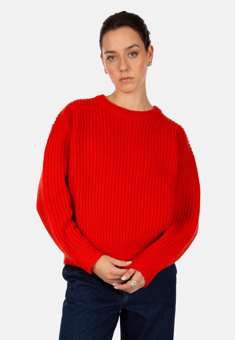 Roter Strickpullover mit rundem Ausschnitt, oversized Ärmeln und gerippter Textur. Kombiniert mit blauen Jeans, die Hände vor sich gefaltet.