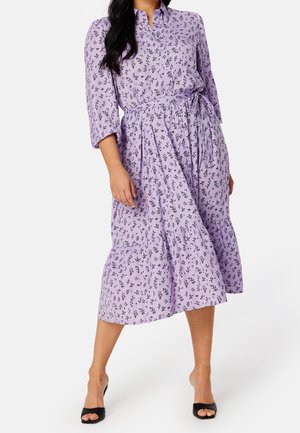 Femme portant une robe midi lavande avec imprimé floral noir, manches trois-quarts, ceinture nouée à la taille, et talons ouverts noirs.