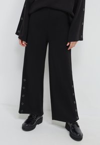 Person in schwarzen, weiten Hosen mit seitlichen Knöpfen und schwarzen, ledernen Schnürschuhen, stehend vor einem schlichten weißen Hintergrund.