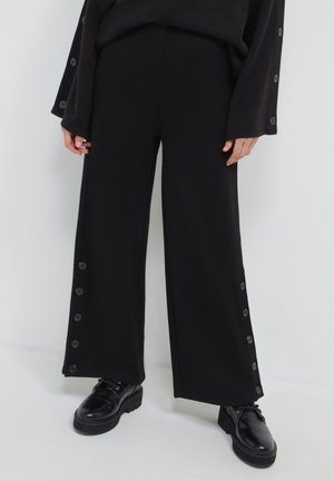 Personne portant un pantalon large noir avec des boutons latéraux et des chaussures en cuir à lacets noires, se tenant devant un fond blanc uni.