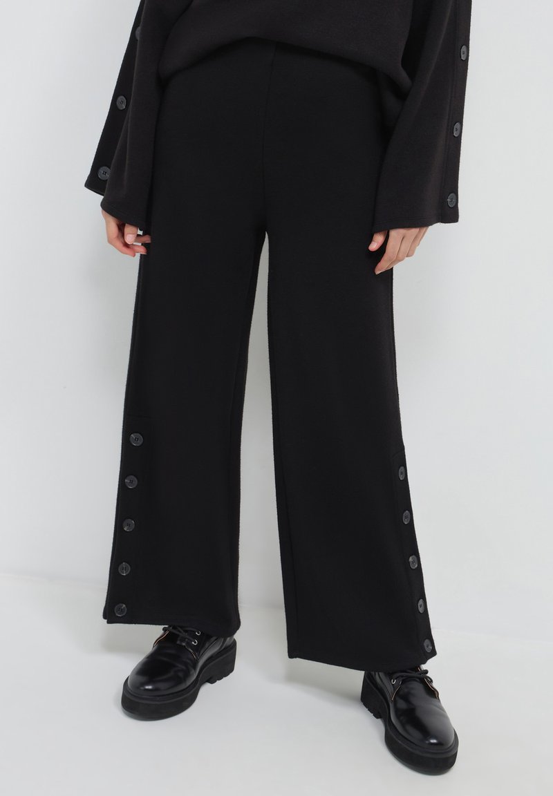 Person in schwarzen, weiten Hosen mit seitlichen Knöpfen und schwarzen, ledernen Schnürschuhen, stehend vor einem schlichten weißen Hintergrund.