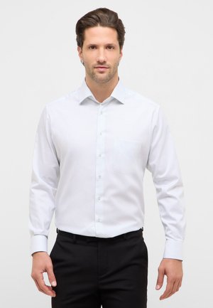 Mand stående med fronten mod kameraet iført en hvid, langærmet skjorte med knapper og sorte bukser foran en ensfarvet baggrund.