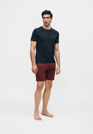 Hombre de pie descalzo, vistiendo una camiseta azul marino de manga corta y pantalones cortos rojo oscuro hasta la rodilla, frente a un fondo claro y liso.