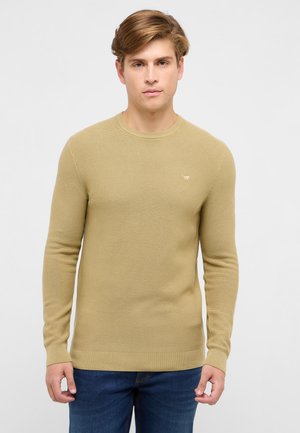 Młody mężczyzna o jasnobrązowych włosach, ubrany w beżowy, długorękawowy sweter z dzianiny i niebieskie dżinsy, stojący na tle jednolitego jasnego tła.