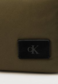 Τσάντα από νάιλον ελιάς με μαύρο ανάγλυφο λογότυπο "cK". Λείο φινίρισμα με διακριτική ριγωτή υφή. Ορθογώνιο σχήμα.