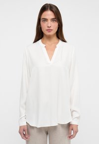 Weiße Bluse mit langen Ärmeln und V-Ausschnitt, aus glattem Stoff; hat einen abgerundeten Saum und subtile Nahtdetails.