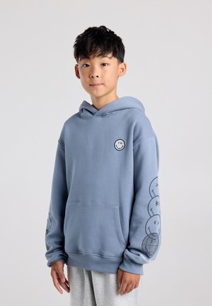 Garçon aux cheveux noirs courts portant un sweat à capuche bleu avec des motifs de visages souriants sur la poitrine et la manche gauche, debout devant un fond clair uni.