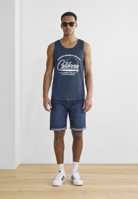 Temno modra brezrokavnica s belim grafičnim tiskom, v kombinaciji z zavihanim denim šortsom; model nosi sončna očala in bele superge.