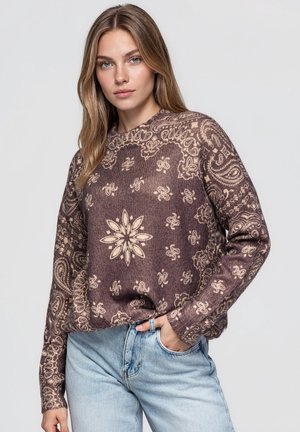 Une femme porte un pull marron à motifs et un jean bleu clair. Elle se tient debout, une main dans sa poche, devant un arrière-plan uni.