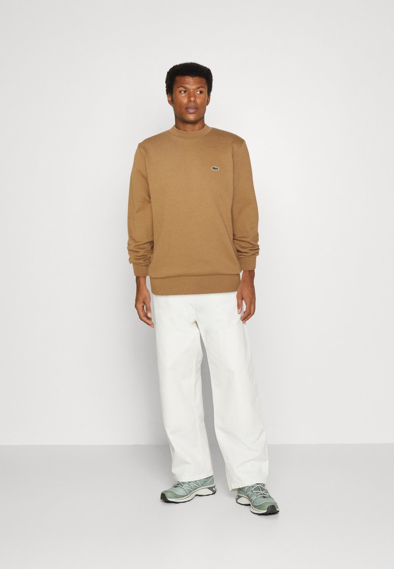 Brauner Hoodie mit gerippten Bündchen und Saum, mit einem kleinen grünen Logo. Kombiniert mit lockeren weißen Hosen und grauen Sneakers.