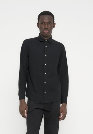 Chemise noire à manches longues avec un col boutonné, dotée de boutons blancs sur le devant et d'une coupe ajustée. Texture unie, sans motifs.