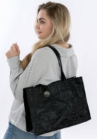 Borsa tote nera in materiale rugoso con due manici neri, dotata di un'etichetta rotonda in argento. Indossata sulla spalla sopra una camicia a righe e jeans blu.
