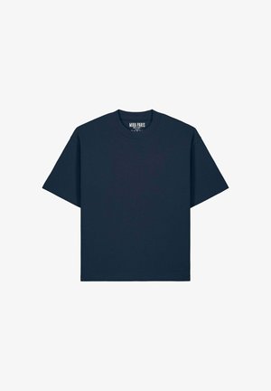 Donna con lunghi capelli intrecciati che indossa una T-shirt oversize blu scuro semplice e pantaloni cargo neri, in piedi contro uno sfondo bianco.