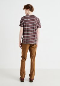 Camiseta de rayas de manga corta en burdeos con rayas crema y azul marino; combinada con pantalones marrones y zapatos oscuros. Vista trasera, fondo minimalista.