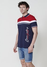 Polo majica u mornarsko plavoj i crvenoj boji s bijelim prugama, s logotipom i grafikom teksta. Uparena s kratkim trapericama s prevrnutim rubom. Materijal od mješavine pamuka.