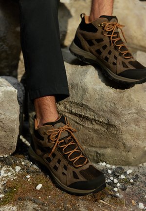 Zapatos de senderismo marrones con parte superior de malla y gamuza, suela de goma y detalles en marrón en los cordones, colocados sobre rocas con fondo de grava y musgo.