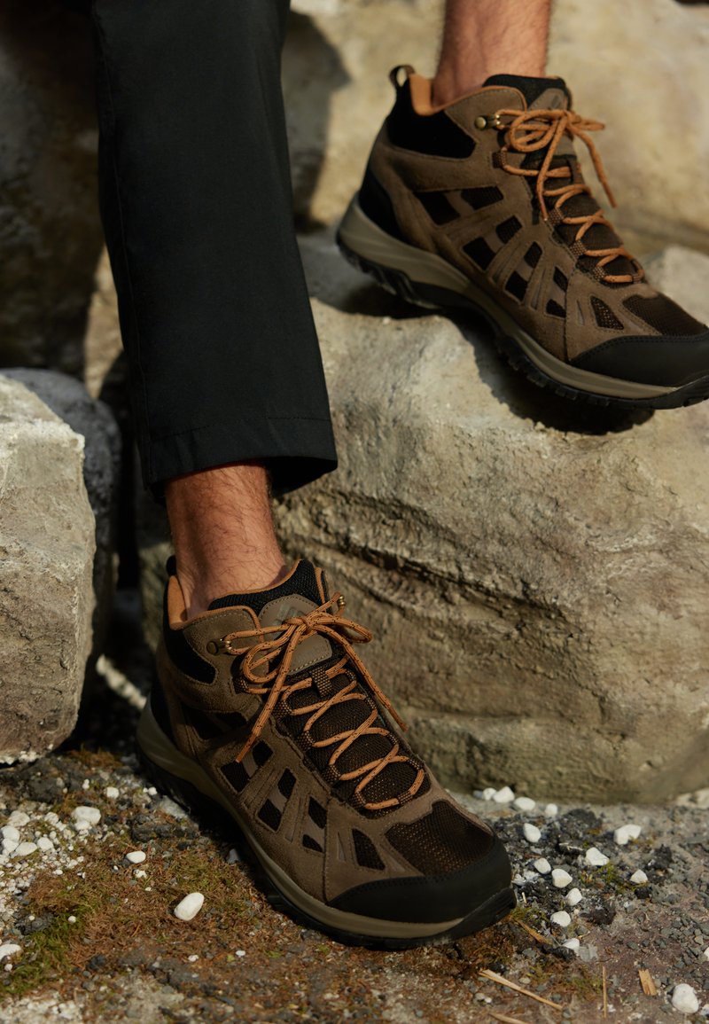 Chaussures de randonnée marron en cuir suédé et mesh, semelle en caoutchouc avec des accents de lacet marron, posées sur des rochers avec un fond de gravier et de mousse.