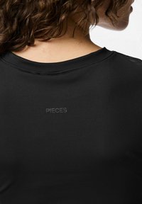 Top nero a maniche corte con una texture liscia. Presenta un colletto rotondo e un sottile logo "PIECES" stampato sul retro.