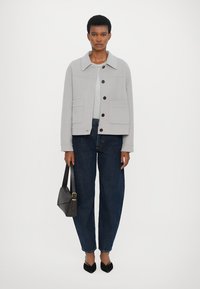 Light grey utility jacket with a collar, front buttons, and two patch pockets. Paired with dark blue relaxed jeans and a brown handbag.