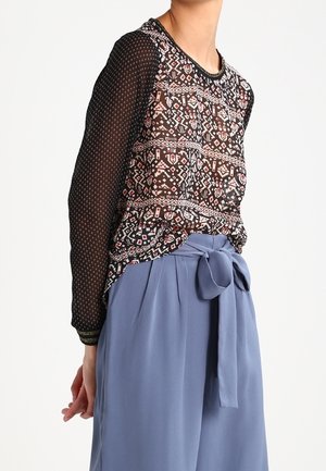 Woman wearing a patterned black sheer long-sleeve blouse tucked into high-waisted loose blue pants with a tied fabric belt.