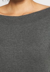 Gros plan sur une personne portant un pull en tricot côtelé gris avec un large décolleté à épaules dénudées mettant en valeur les clavicules.